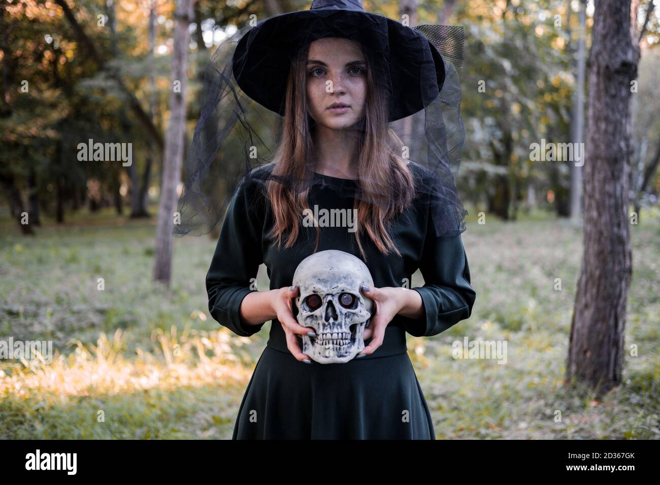 Jeune belle femme mignonne en robe sombre et chapeau de sorcière tient un crâne dans ses mains. Costume de fête d'Halloween. Forêt, parc avec arbres d'automne Banque D'Images