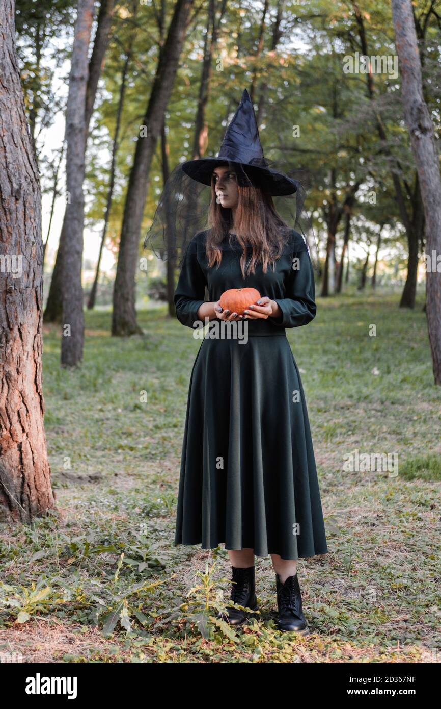 Jeune mignonne belle femme en robe sombre et chapeau de sorcière tient une citrouille orange dans ses mains. Costume de fête d'Halloween. Forêt, parc avec arbres d'automne Banque D'Images