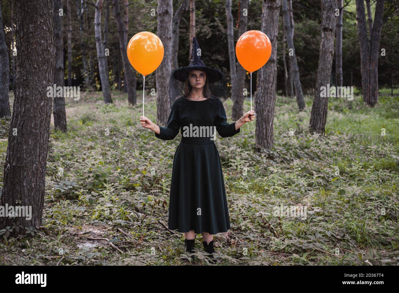 Jeune mignonne belle femme en robe sombre et chapeau de sorcière tient des ballons d'orange dans ses mains. Costume de fête d'Halloween. Forêt, parc avec arbres d'automne Banque D'Images