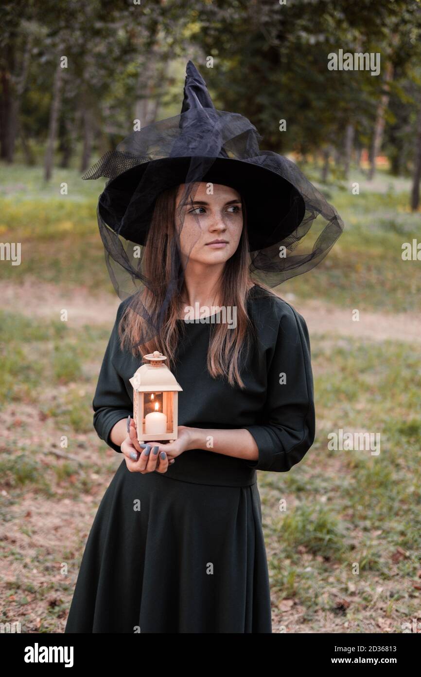 Une jeune femme en robe sombre et chapeau de sorcière tient une lanterne avec des bougies dans ses mains et illumine la forêt. Costume de fête d'Halloween. Garez-vous avec l'autom Banque D'Images