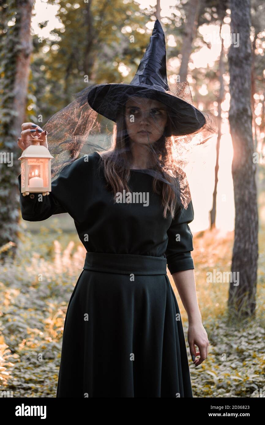 Une jeune femme en robe sombre et chapeau de sorcière tient une lanterne avec des bougies dans ses mains et illumine la forêt. Costume de fête d'Halloween. Garez-vous avec l'autom Banque D'Images