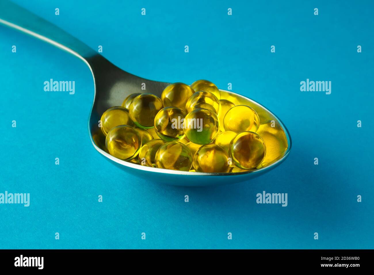 Tas de capsules rondes d'huile de poisson dans une cuillère métallique sur fond bleu, mise au point sélective, gros plan Banque D'Images