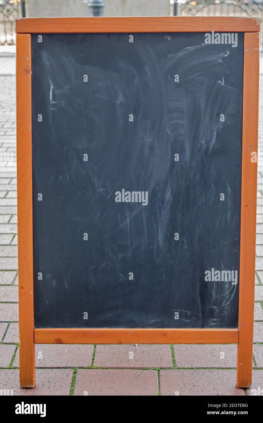 Tableau noir avec des traces de craie dans un cadre en bois marron sur une rue de carreaux beiges sur un fond flou de clôture. Maquette pour la conception de menus. Banque D'Images
