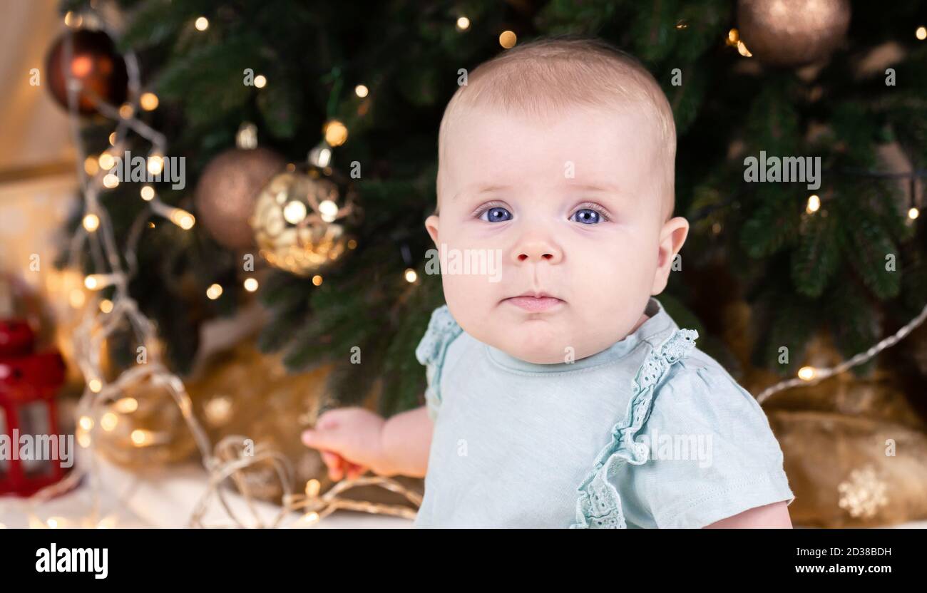 Bébé de Noël regardant l'appareil photo. Une petite fille mignonne dans une robe bleue et un chapeau blanc exprime ses émotions. Concept de Noël avec petit enfant, arbre et Banque D'Images