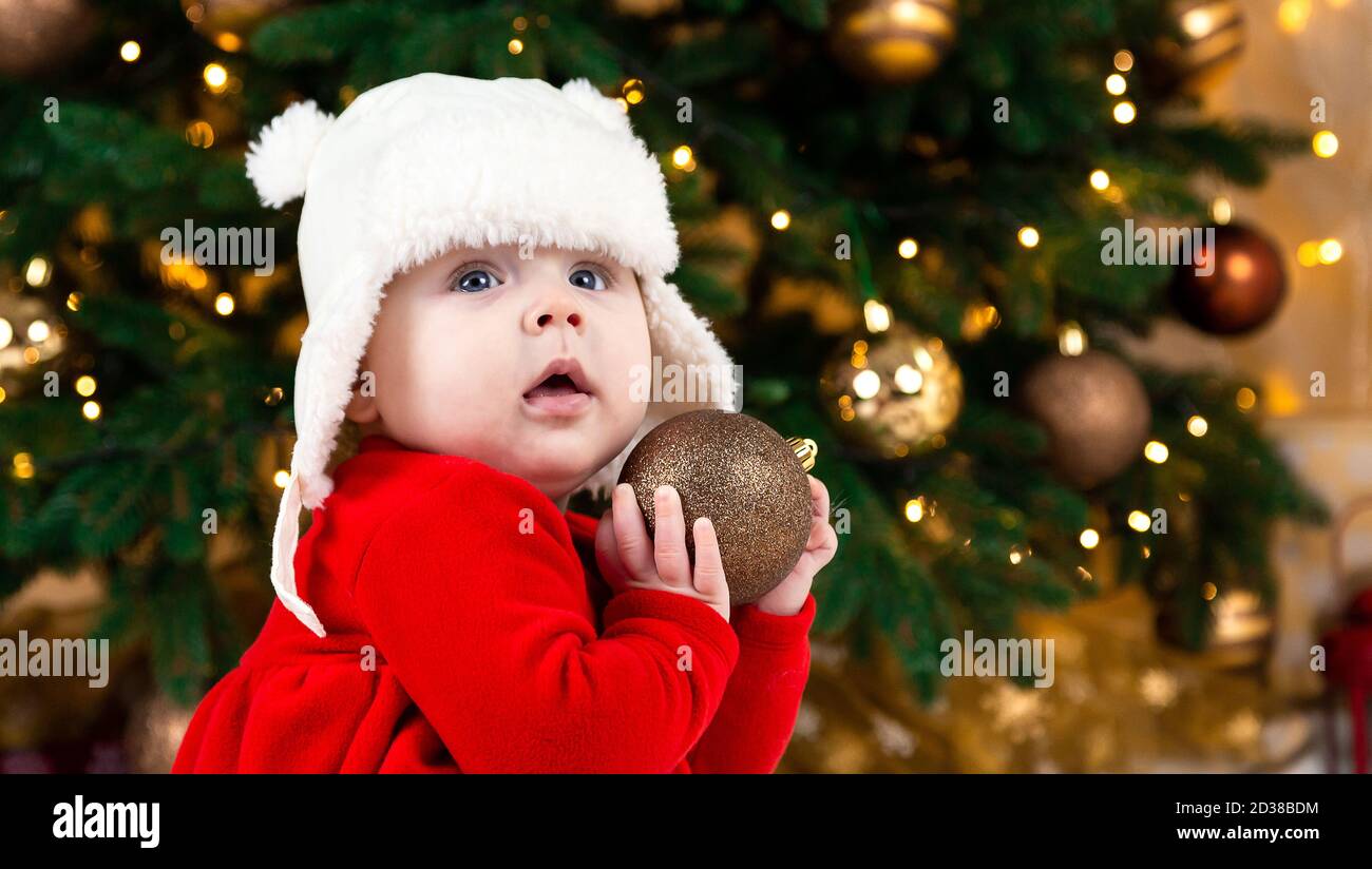 Boule De Noel Pour Bebe Une Petite Fille Mignonne Veque D Une Robe Rouge Et D Un Chapeau Blanc Exprime Ses Emotions Concept De Noel Avec Petit Enfant Arbre Et Guirlande Sur B Photo