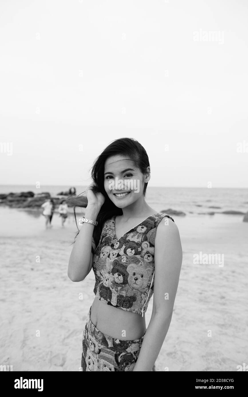 Young happy Asian woman smiling tout en fixant ses cheveux à la plage publique de Hua Hin en Thaïlande Banque D'Images