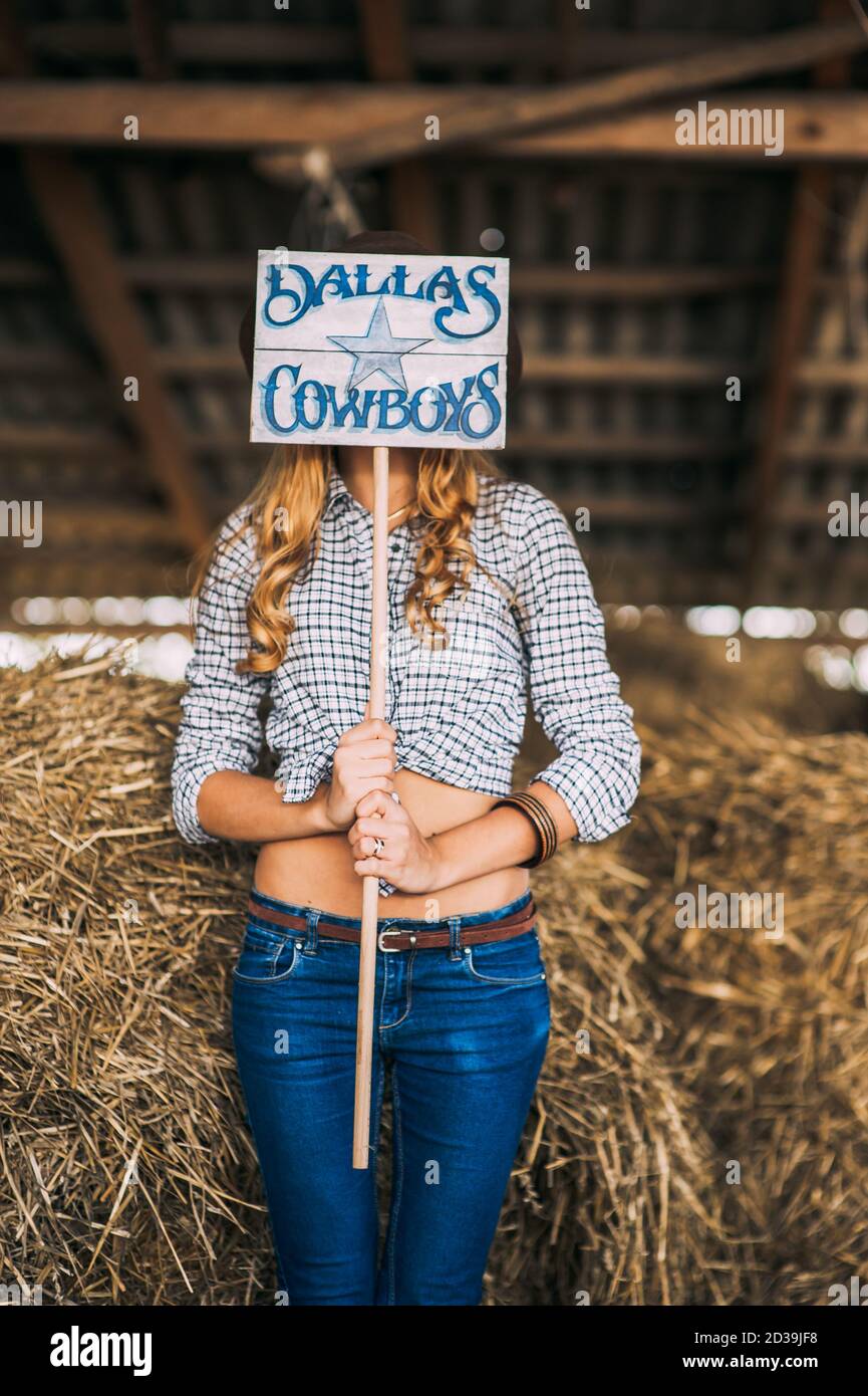 Femme blonde sans visage en vêtements de cow-boy tenant une planche en bois avec l'inscription dallas Cowboys devant son visage sur un fond de foin. Rustique Banque D'Images
