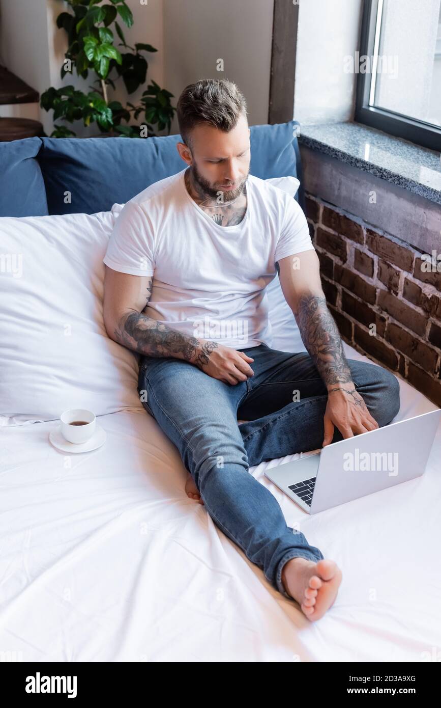 jeune journaliste indépendant tatoué en jean et t-shirt blanc utilisant un ordinateur portable dans le lit près d'une tasse de café Banque D'Images