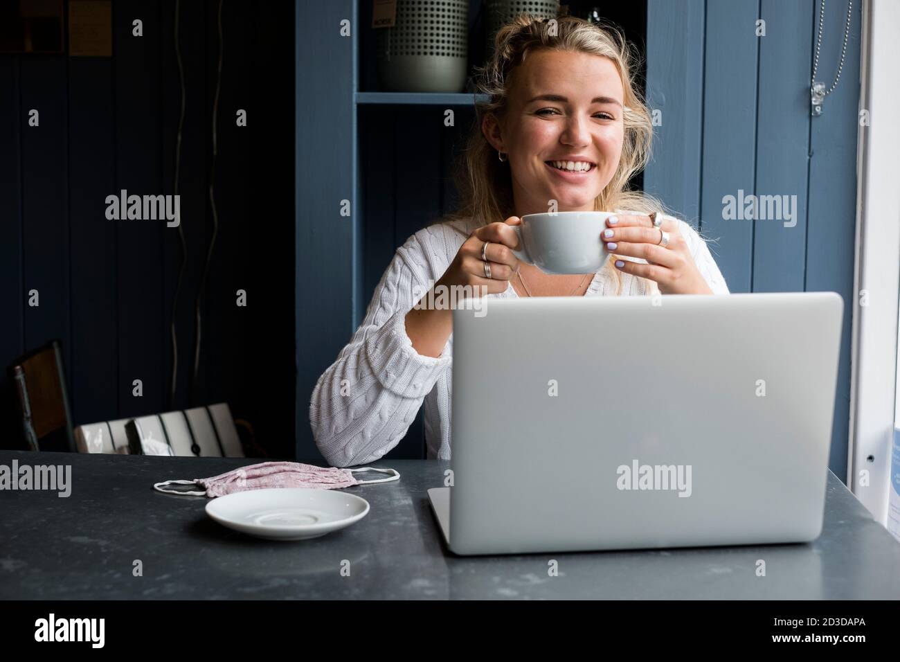 Jeune femme blonde assise seule à une table de café avec un ordinateur portable et une tasse de café Banque D'Images
