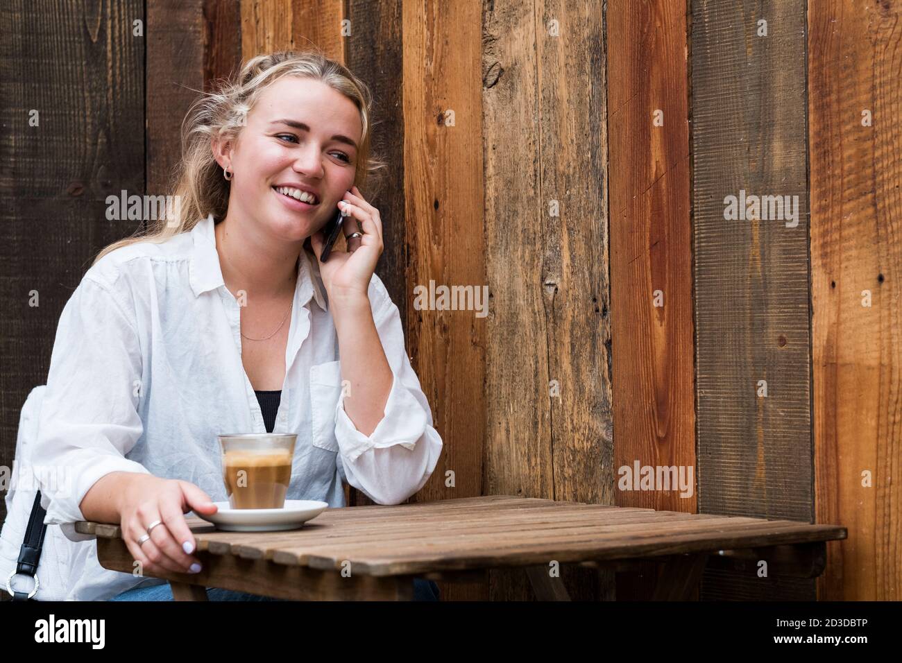 Jeune femme blonde assise seule dans un café, utilisant un téléphone mobile, travaillant à distance. Banque D'Images