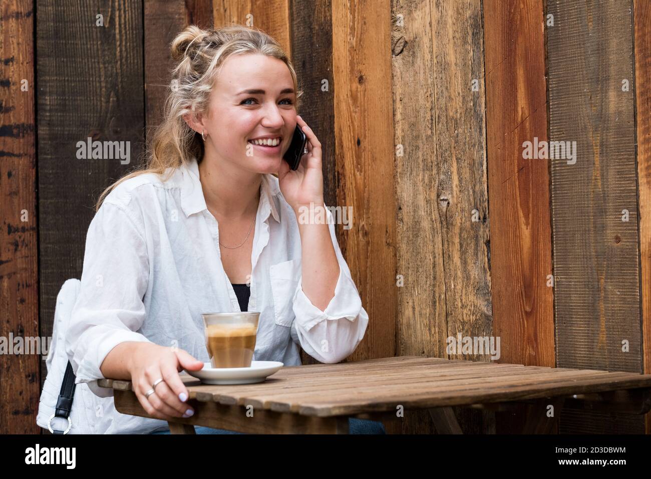 Jeune femme blonde assise seule dans un café, utilisant un téléphone mobile, travaillant à distance. Banque D'Images