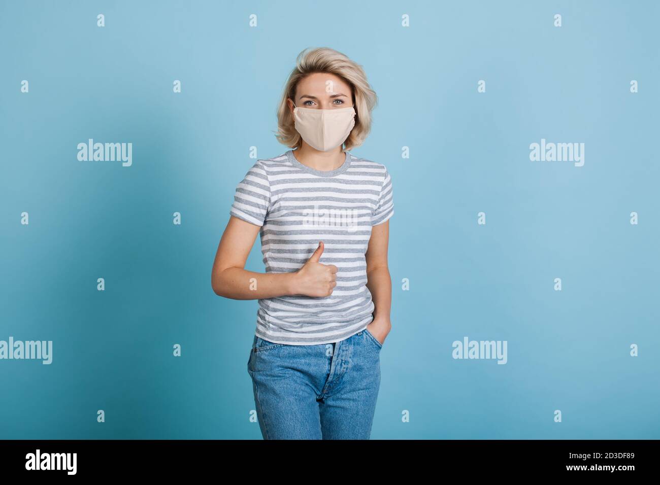 Jolie femme blonde avec masque médical sur le visage est gestante le signe similaire sur un mur bleu de studio Banque D'Images