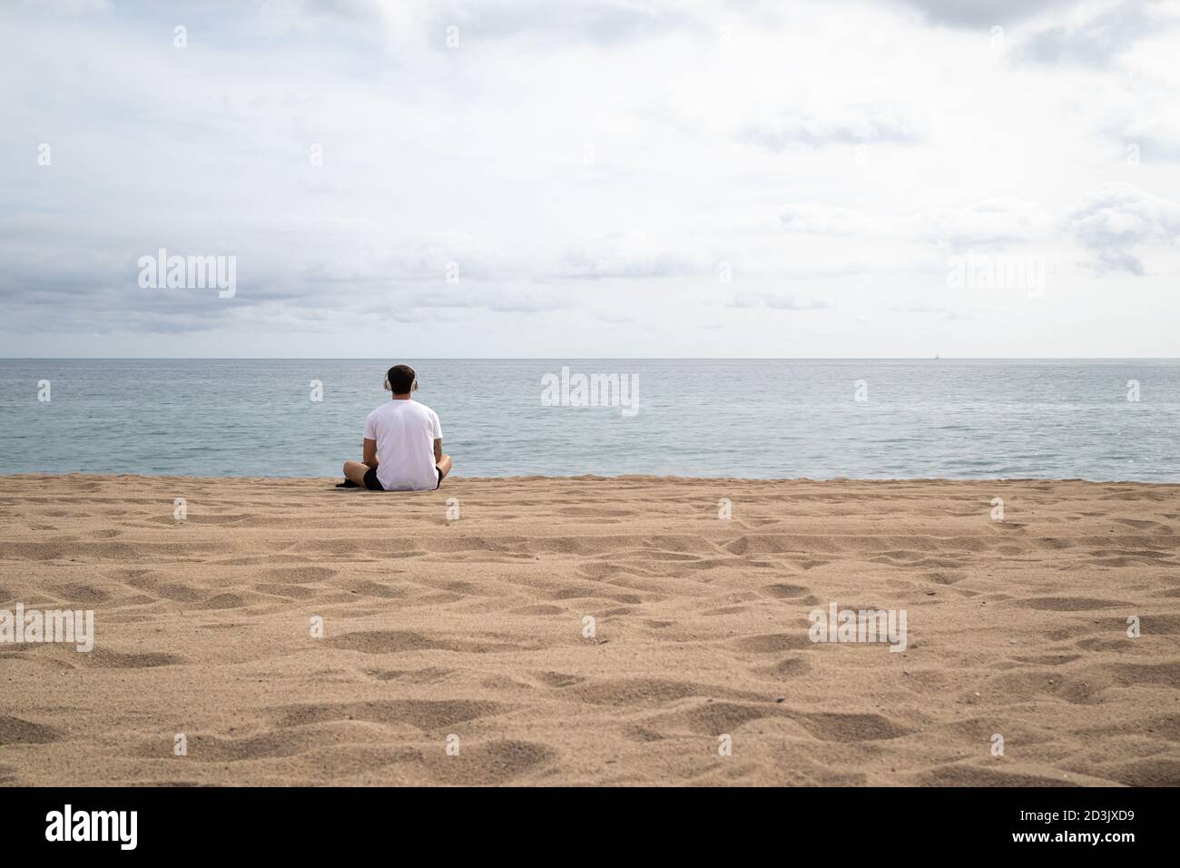 Jeune homme méditant dans des écouteurs assis sur la plage de sable par la mer en jour nuageux Banque D'Images