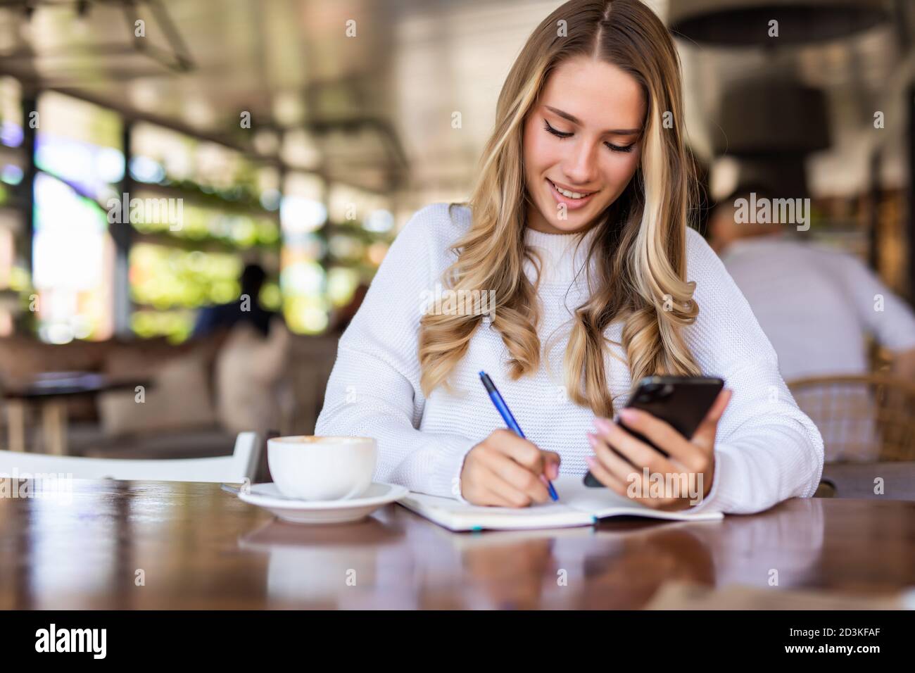Jeune femme lisant les nouvelles des réseaux sociaux en utilisant le téléphone mobile au café. Faire des recherches en ligne sur smartphone tout en créant un article pour le partage d'écriture Banque D'Images