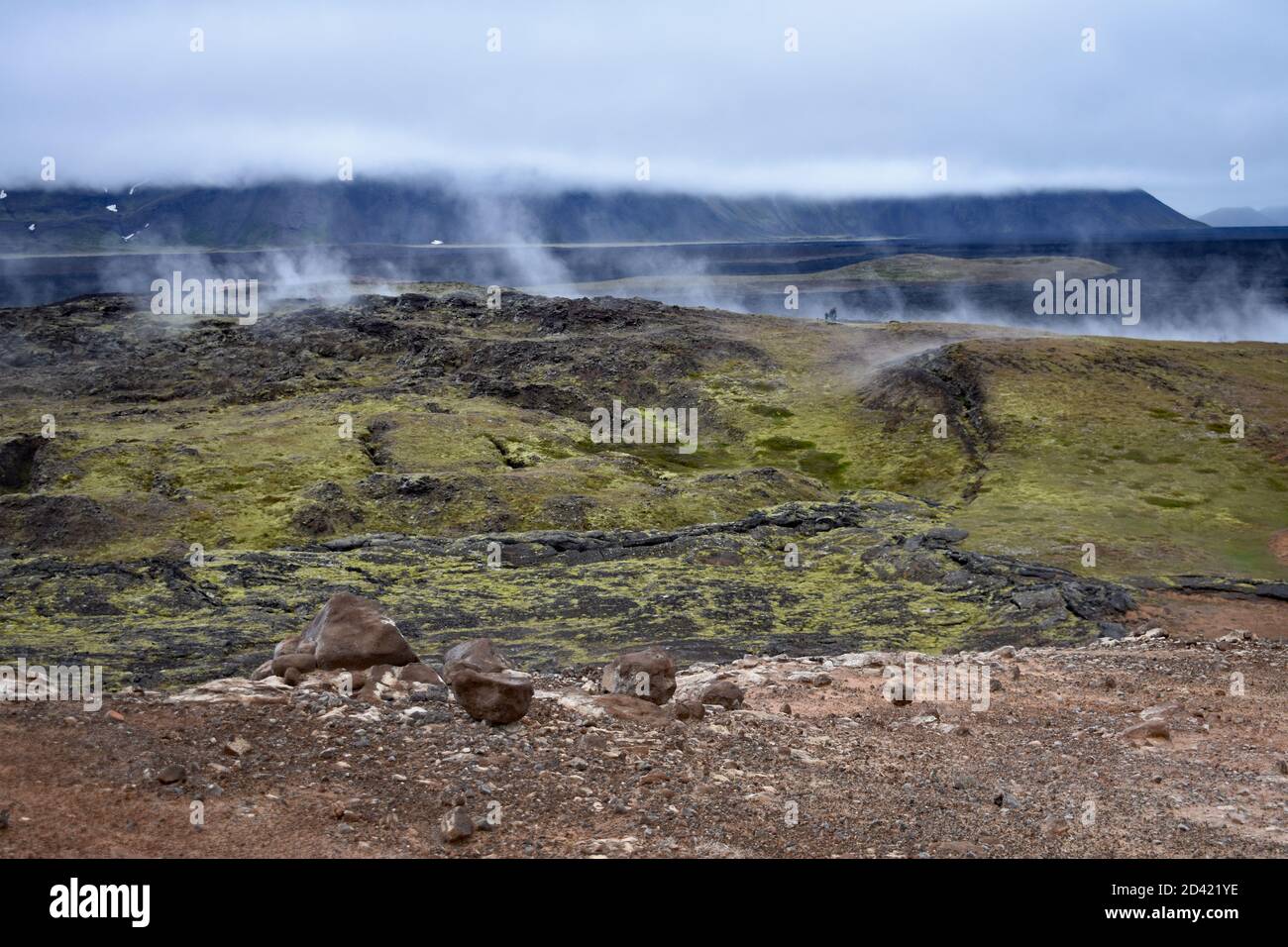 La vapeur s'élève dans une ligne dans les champs de Krafla Lava, dans la région du lac Myvatn, dans le nord de l'Islande. Les nuages couvrent partiellement les montagnes derrière la roche volcanique. Banque D'Images