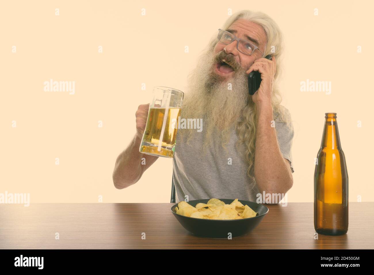 Heureux homme barbu senior parlant au téléphone et tenant verre de bière avec bol de chips de pomme de terre sur bois tableau Banque D'Images