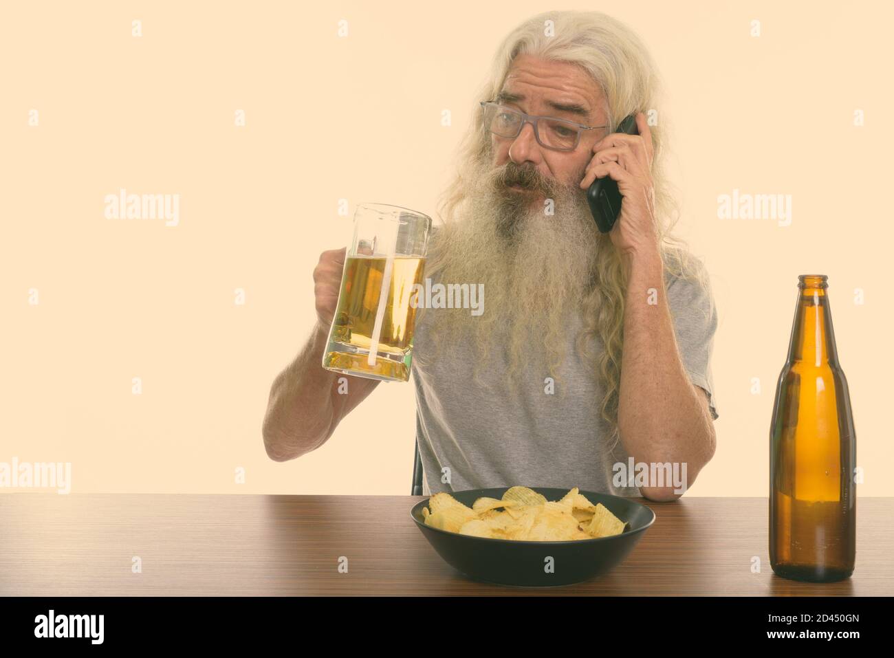 Studio shot of senior bearded man talking on mobile phone en maintenant le verre de bière avec bol de croustilles de pommes de terre sur la table en bois Banque D'Images