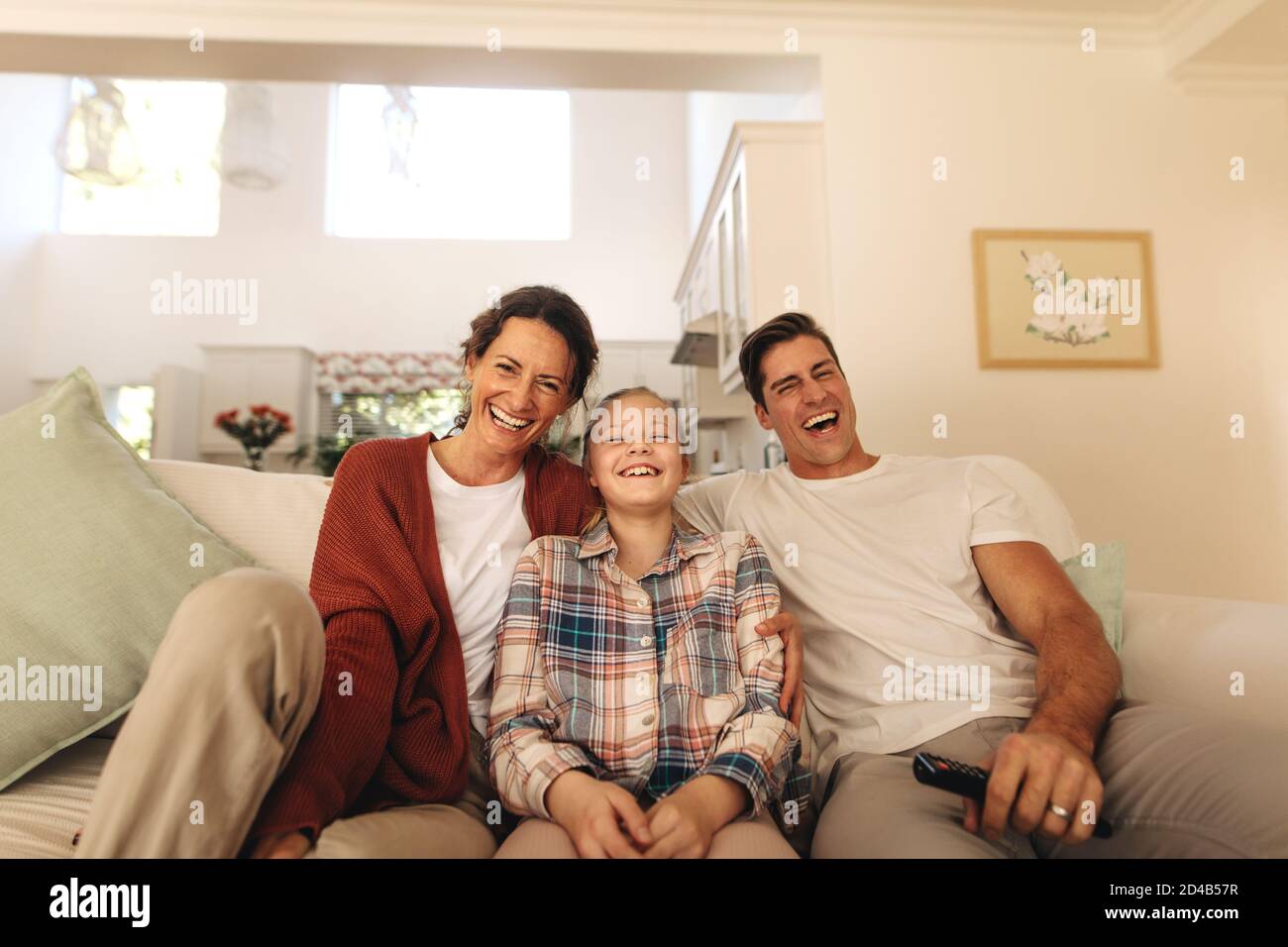 Une famille souriante assise sur un canapé tout en regardant la télévision à la maison. Famille de trois personnes se détendant à la maison en regardant la télévision. Banque D'Images