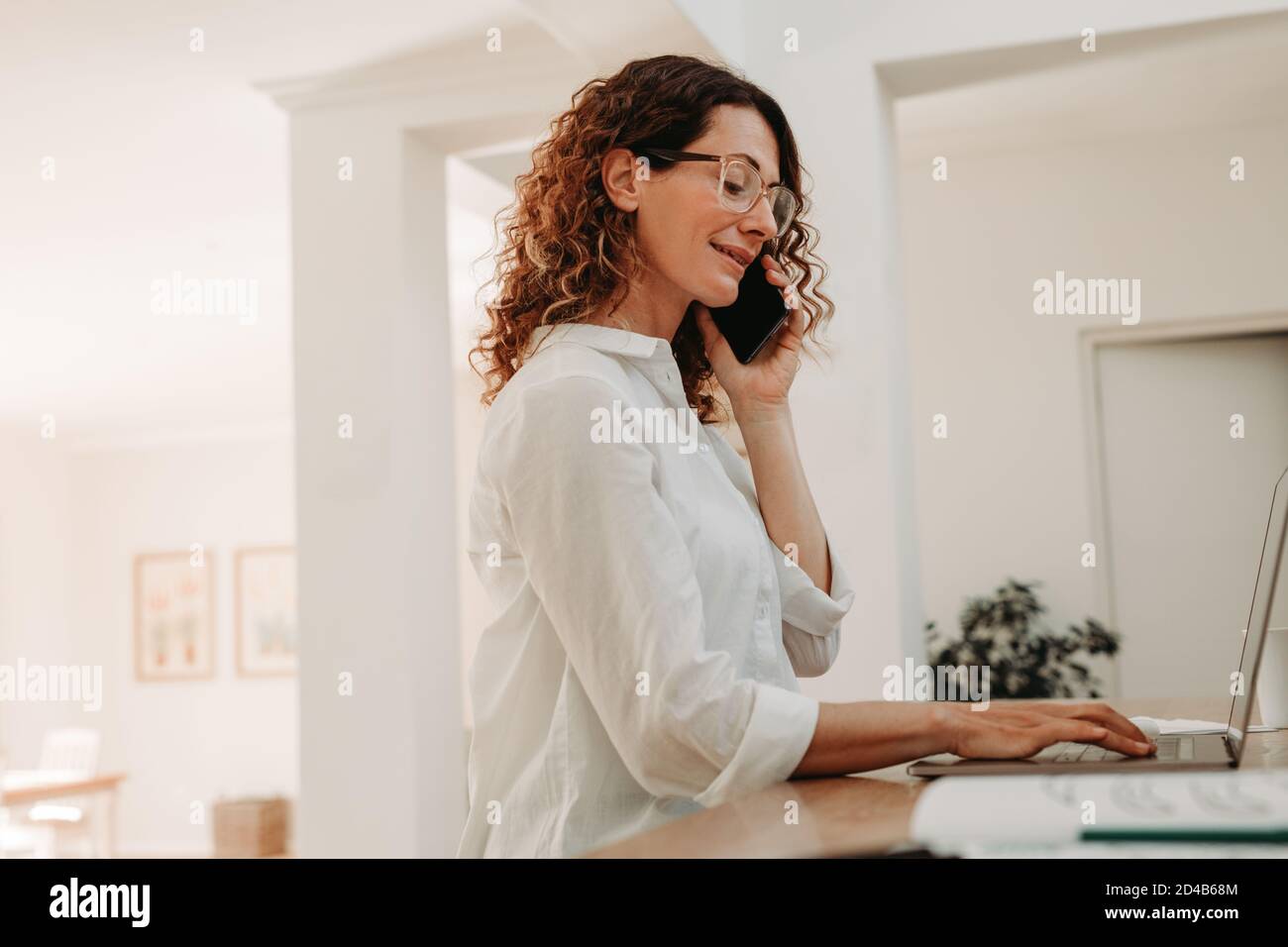 Femme heureuse travaillant sur ordinateur portable et parlant sur téléphone portable à la maison. femme d'affaires assise à table et travaillant. Banque D'Images