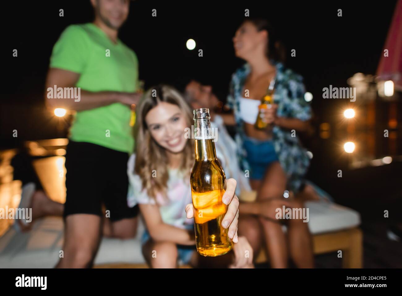 Objectif sélectif de la femme montrant une bouteille de bière près de l'ami et amis à l'extérieur la nuit Banque D'Images