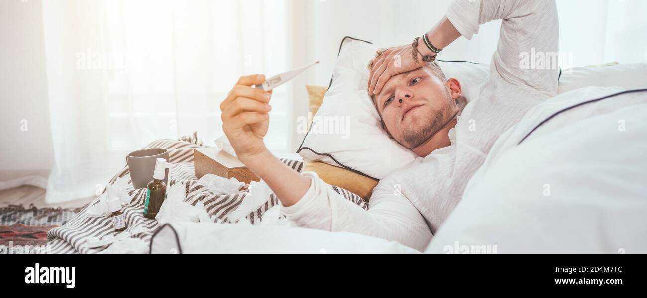 Malsain et fatigué regardant un thermomètre médical comme étant allongé dans un lit confortable à la maison à côté d'un grand nombre de mouchoirs en papier et de médicaments utilisés sur la couverture. Mers Banque D'Images