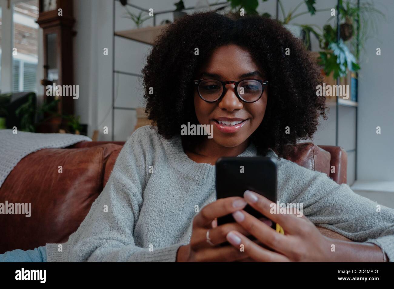 Femme de race mixte portant des lunettes souriantes défilant sur un smartphone assis sur un canapé dans un salon minimaliste. Banque D'Images