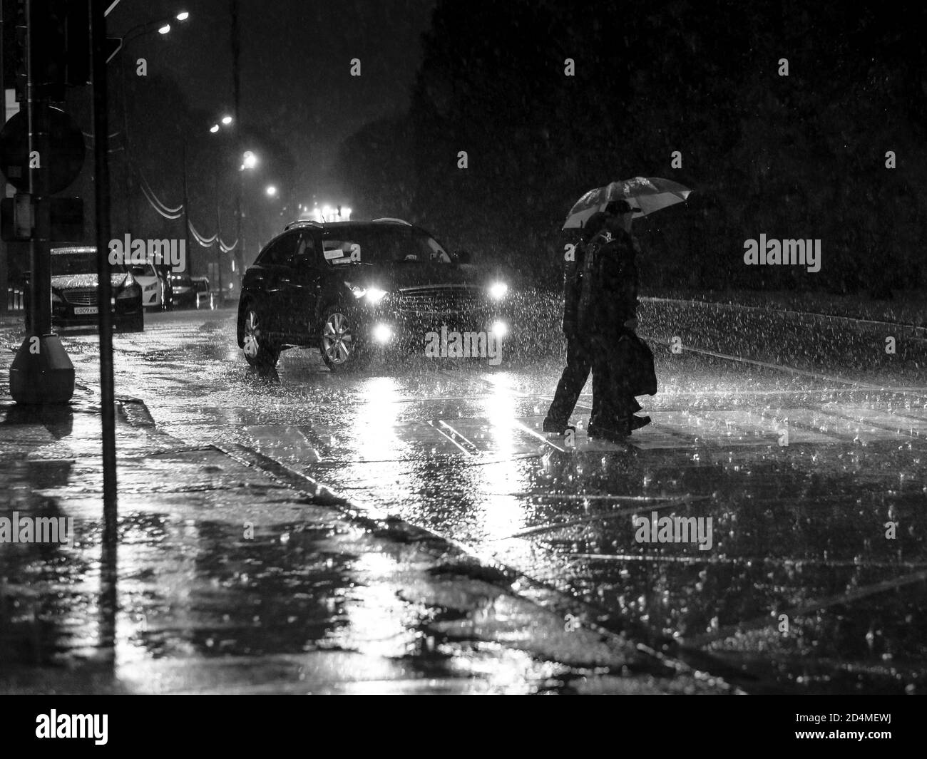 Vue sur une traversée piétonne de la ville la nuit pendant une lourde averse. Silhouettes de personnes avec parasols dans les phares de la voiture. Personnes Banque D'Images