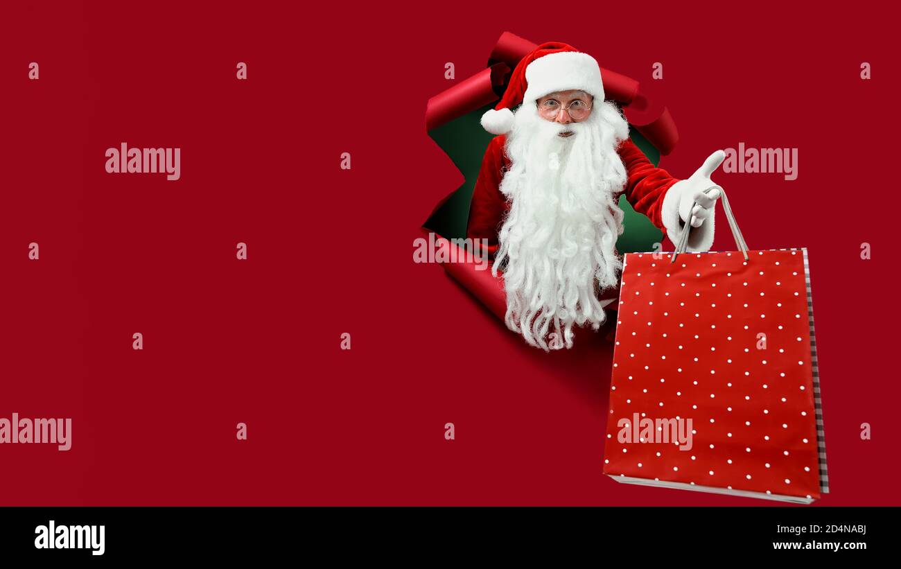 Le père Noël tient le paquet de magasins dans la main par un trou de papier. Homme barbu dans chapeau de père noël regardant à travers trou sur papier rouge. Banque D'Images