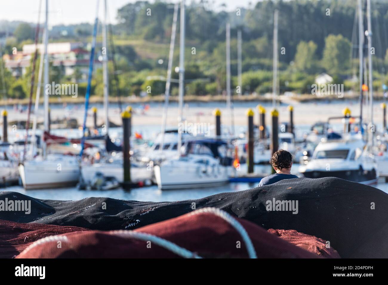 Un homme d'âge moyen non identifiable lit un livre parmi les filets de pêche avec de petits bateaux à voile amarrés dans un club de yacht en Galice en arrière-plan. SH Banque D'Images