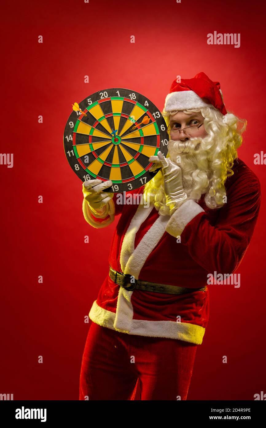 L'acteur masculin dans un costume du Père Noël tient un fléchettes cible dans ses mains et pose sur une obscurité arrière-plan rouge Banque D'Images