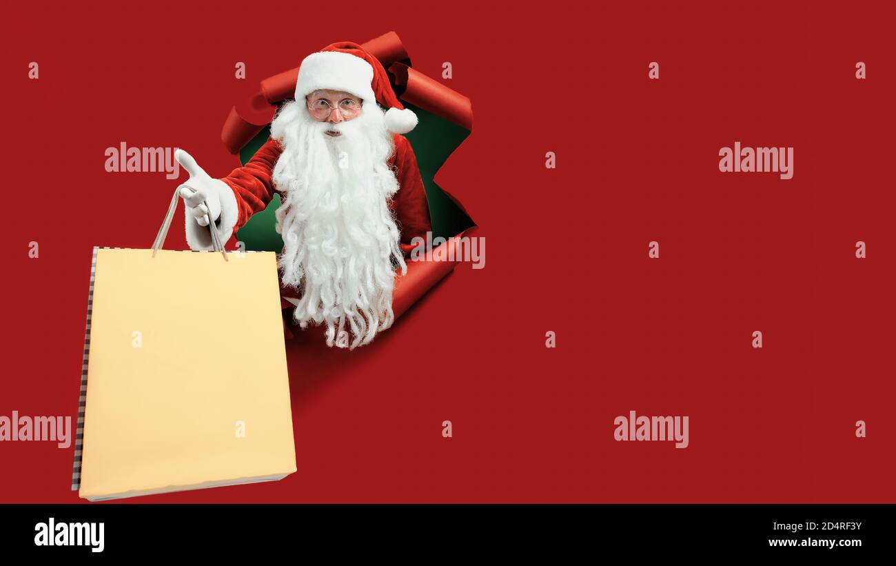 Le père Noël tient le paquet de magasins dans la main par un trou de papier. Homme barbu dans chapeau de père noël regardant à travers trou sur papier rouge. Banque D'Images