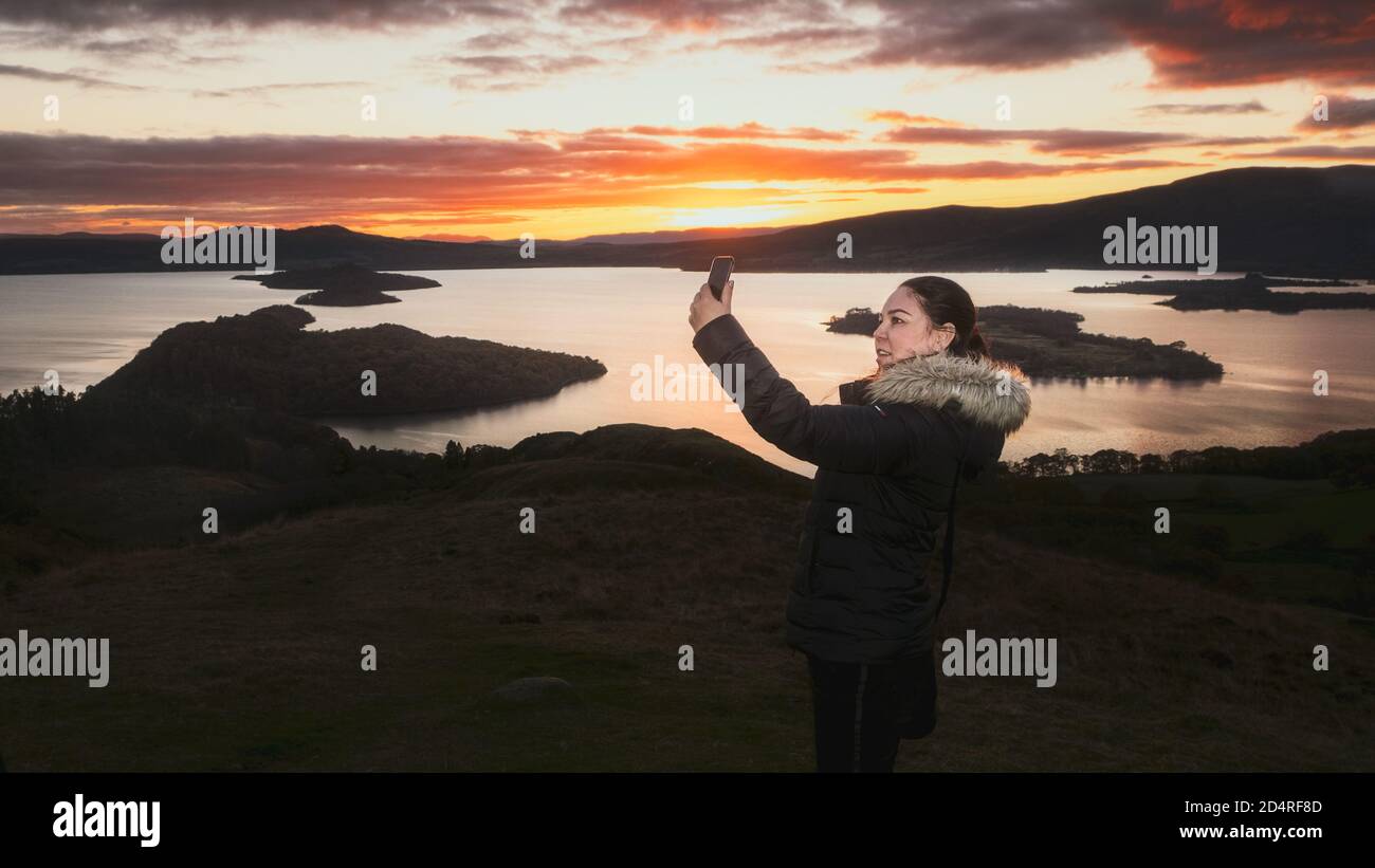 Une femme faisant un selfie à partir d'un smartphone se tient sur une montagne au bord du lac, sur fond d'un ciel de coucher de soleil brûlant. Conic Hill, Écosse, United Banque D'Images