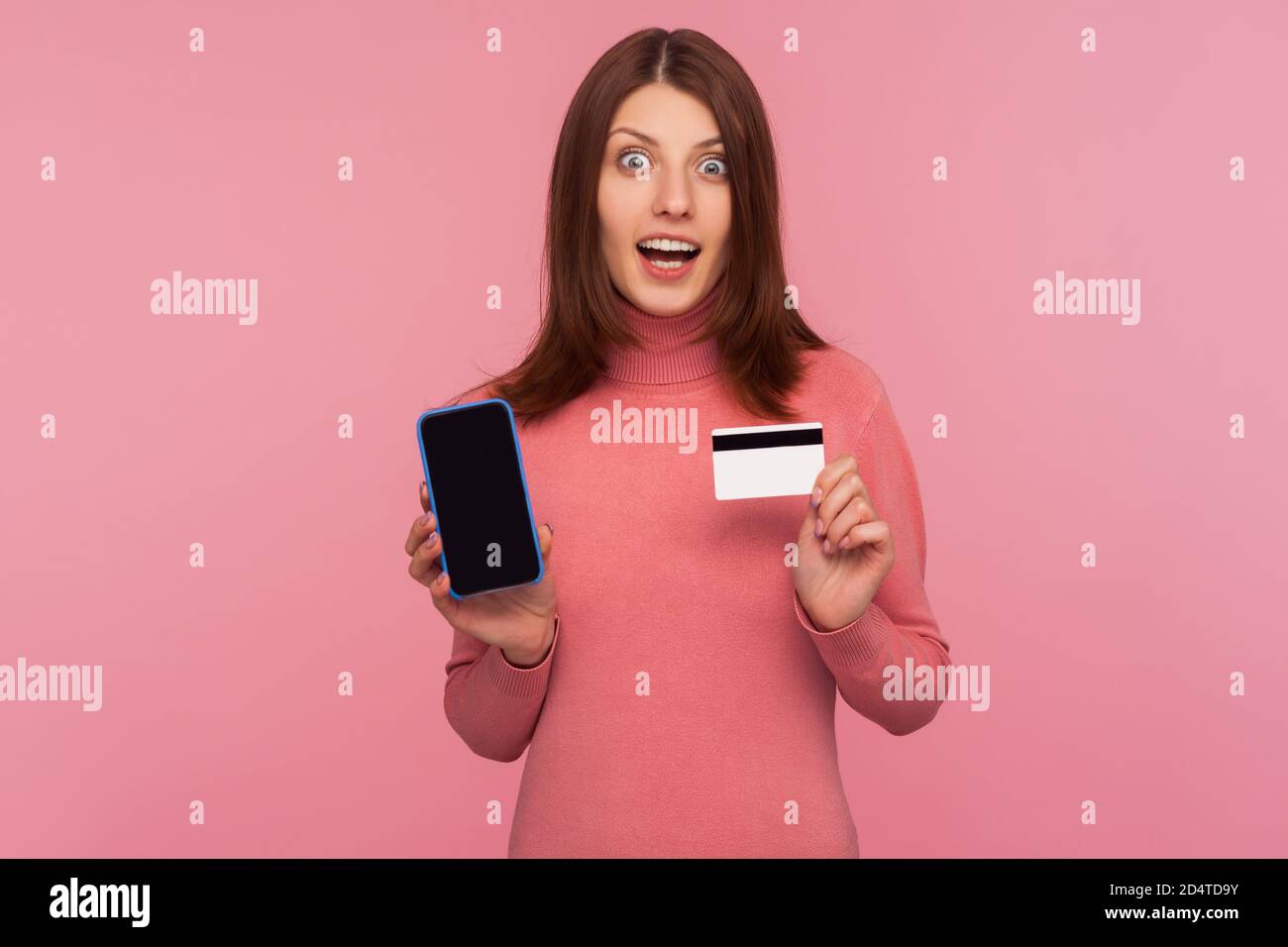 Femme brunette stupéfiée en chandail rose montrant téléphone cellulaire et carte de crédit, regardant la caméra avec l'expression choquée, surprisée avec les banques en ligne. Po Banque D'Images