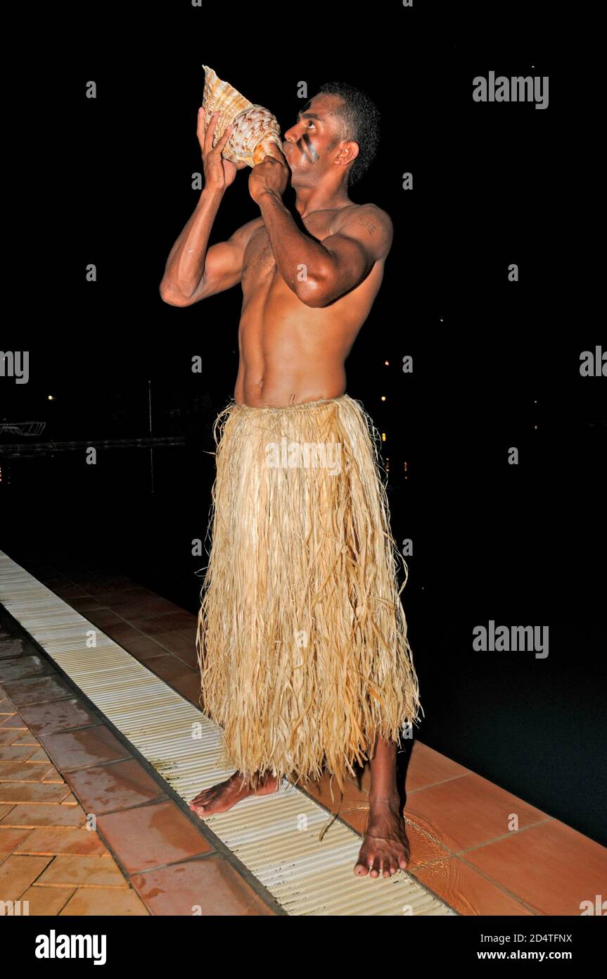 Un jeune Fijian portant un Sulu (jupe en herbe) Souffle dans un seashell appelé un Davui pour invoquer le communauté du village pour assister à une cérémonie à laquelle assistera le Banque D'Images