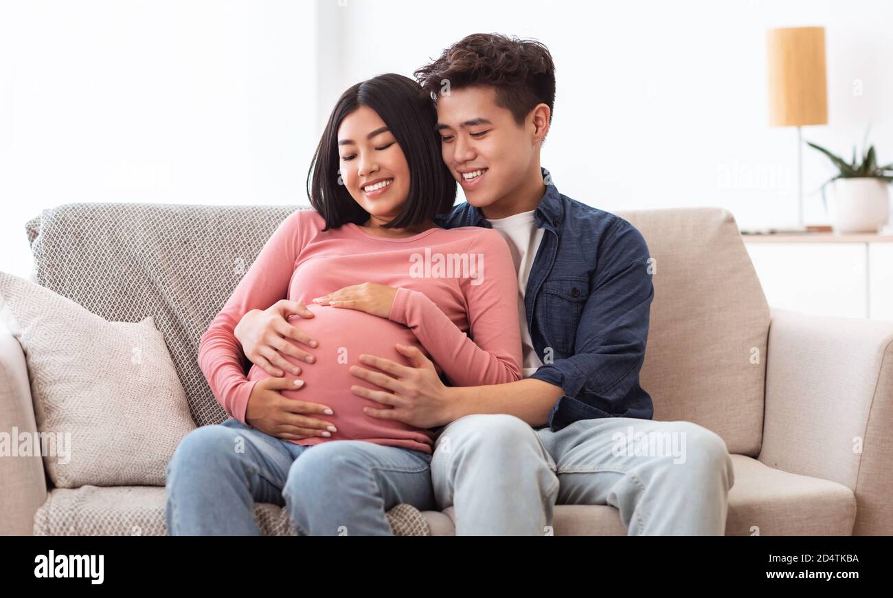 Heureux mari asiatique embrassant femme enceinte assis ensemble à la maison Banque D'Images