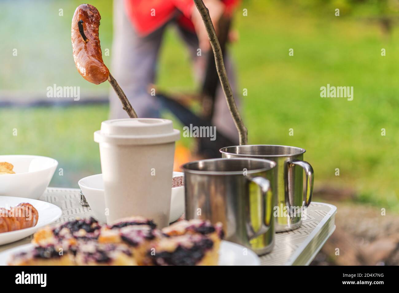 Table avec tasses à café en métal et gâteaux. Polir la saucisse sur un bâton. Camping avec Campfire Made Food Banque D'Images