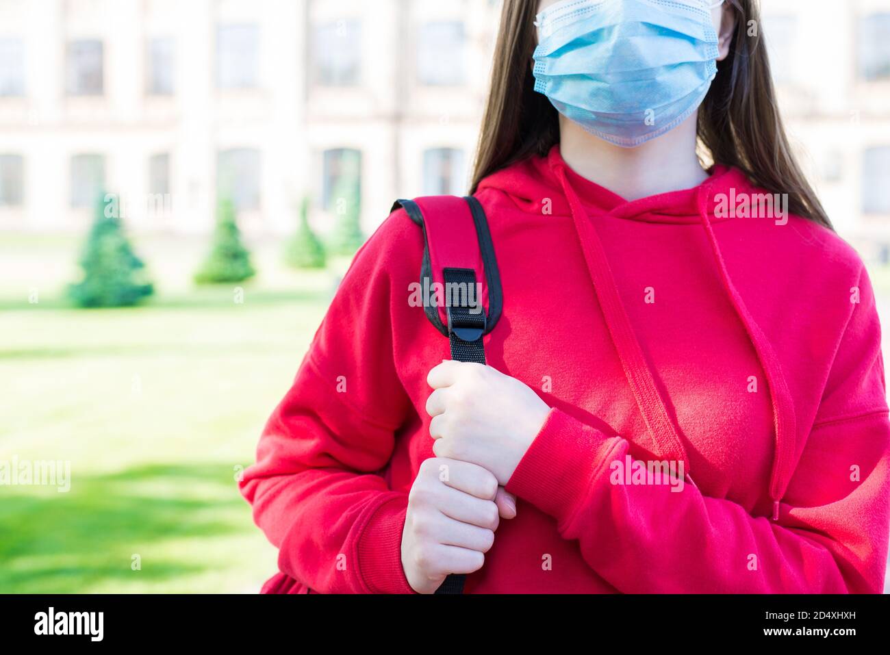 La vie normale en utilisant le concept de masque facial. Petit gros plan portrait photo de fille sérieuse en chandail rouge décontracté et masque filtrant debout près de construction d'école Banque D'Images