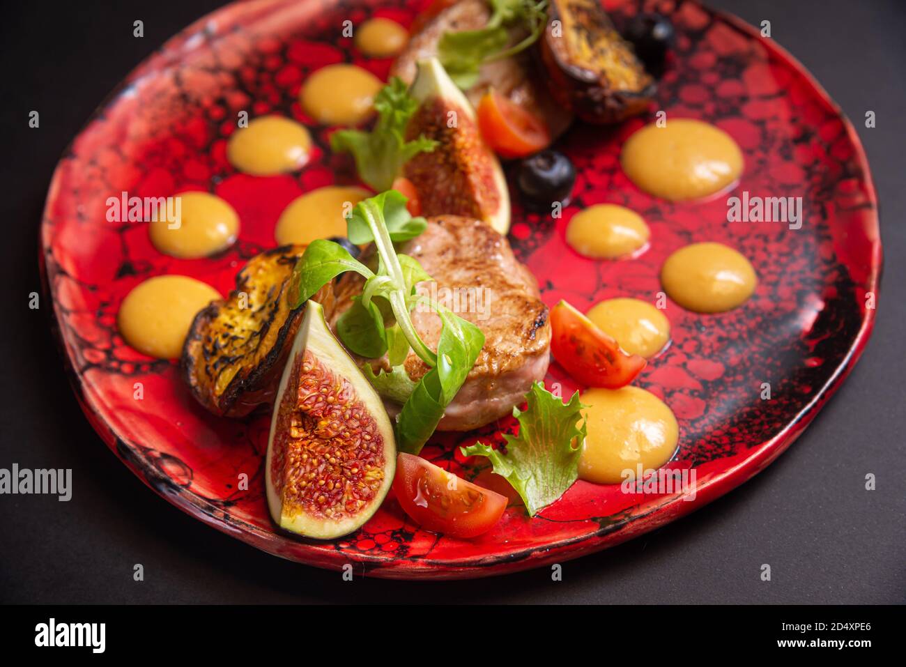 Salade chaude, porc frit, tranches de figues, pêches frites, bleuets, tranches de tomates cerises, feuilles vertes, sauce à la purée de citrouille. Sur une assiette rouge faite à la main Banque D'Images