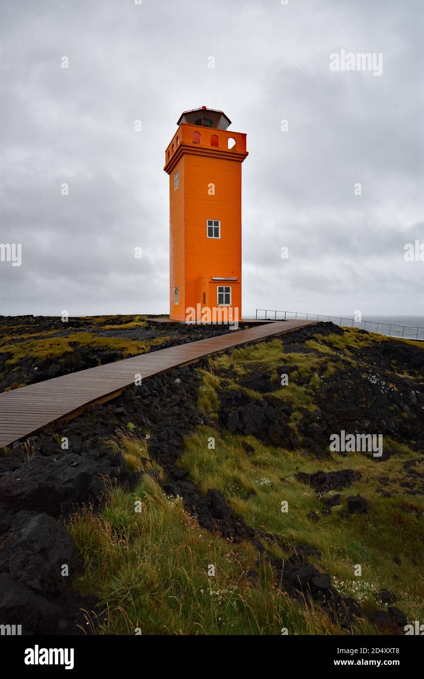 Un chemin en bois coupe une roche de lave noire recouverte de mousse et d'herbe jusqu'au phare orange de Svortuloft sur la péninsule de Snaefellsnes, dans l'ouest de l'Islande. Banque D'Images