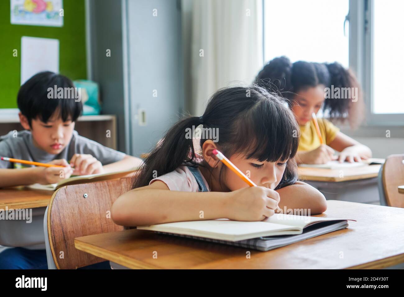 Fille d'école assis à l'école écrire dans le livre avec crayon, étudier, éducation, apprendre. Enfants asiatiques dans la classe. Diversité des élèves. Banque D'Images