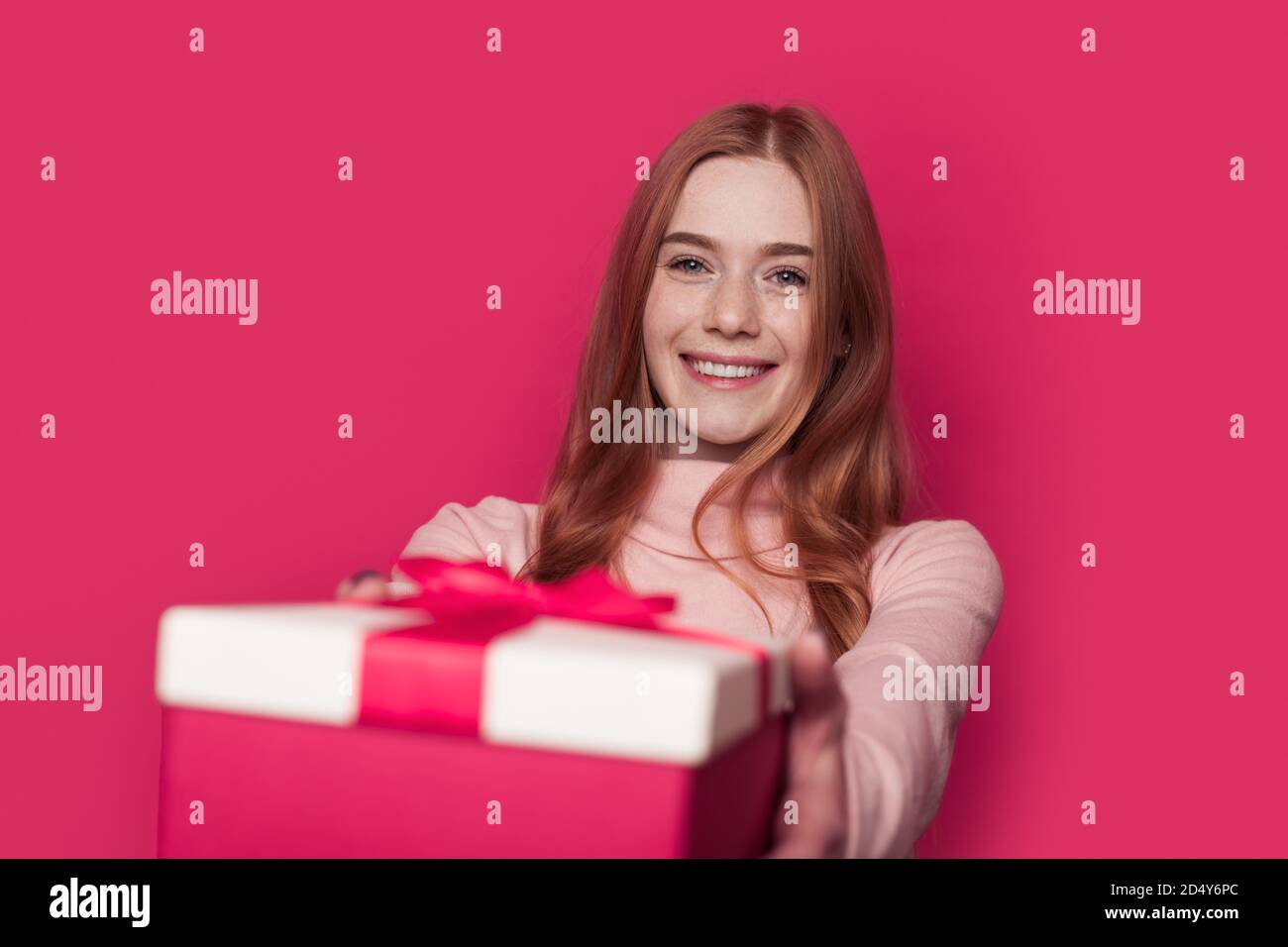 Charmante femme au gingembre avec des taches de rousseur donne un cadeau à l'appareil photo souriant sur un mur rose au studio Banque D'Images