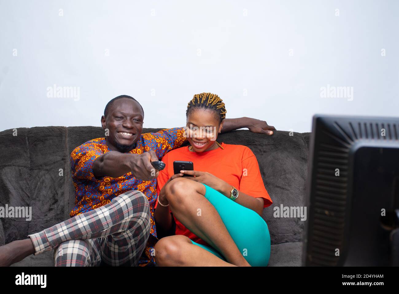 couple africain assis ensemble sur un canapé, l'homme regardant la télévision et la femme utilisant son téléphone, les deux souriants Banque D'Images