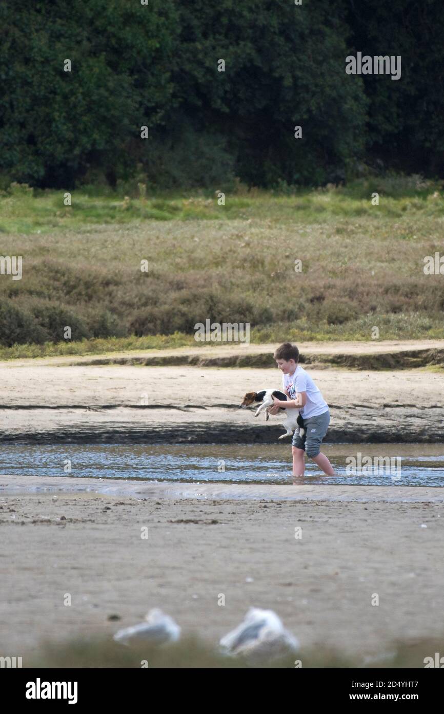 Un jeune garçon marche pieds nus et porte son petit chien à marée basse sur la rivière Gannel à Newquay, dans les Cornouailles. Banque D'Images