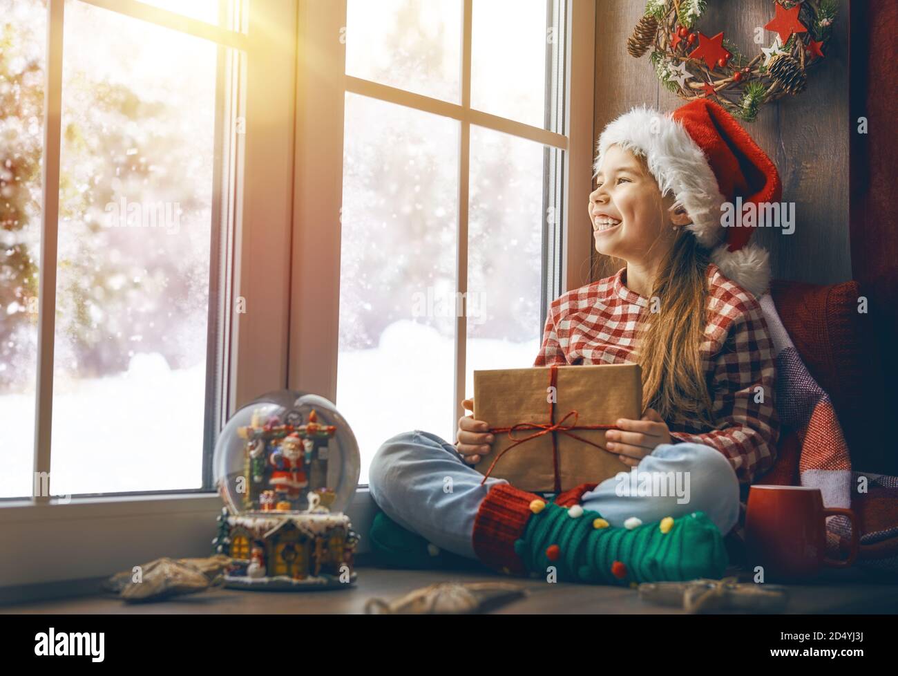 Joyeux Noël et joyeuses fêtes! Petite fille mignonne assise près de la fenêtre avec un coffret cadeau et regardant la forêt d'hiver. Chambre décorée. K Banque D'Images