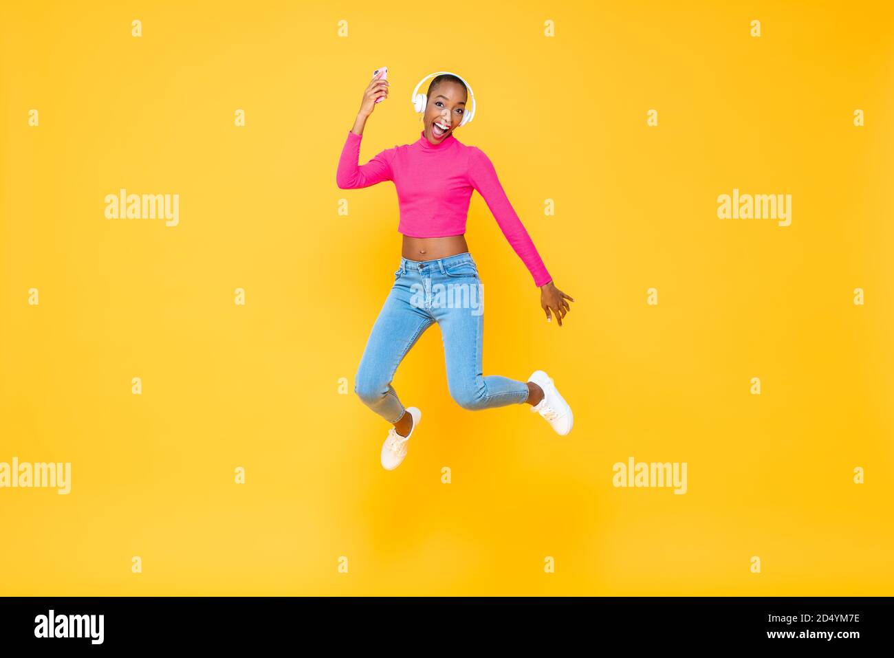 Portrait complet du saut heureux énergique Afro-américaine femme porter un casque pour écouter de la musique à partir d'un smartphone isolé des couleurs c'est tout Banque D'Images