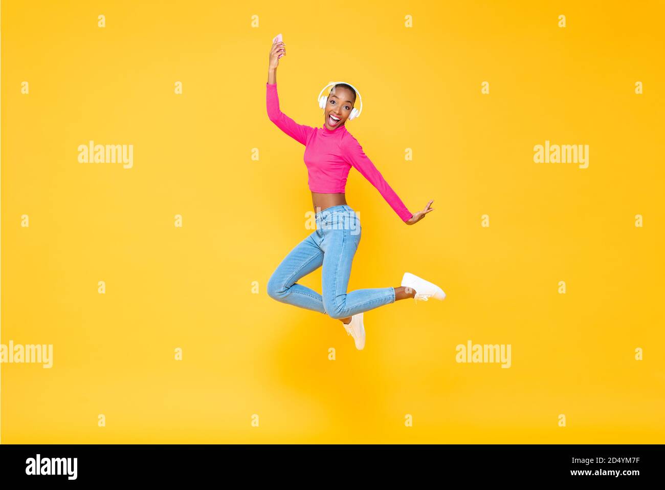 Portrait amusant d'une femme afro-américaine énergique souriante portant un casque écouter de la musique depuis un smartphone et sauter à l'écart des couleurs jaune Banque D'Images
