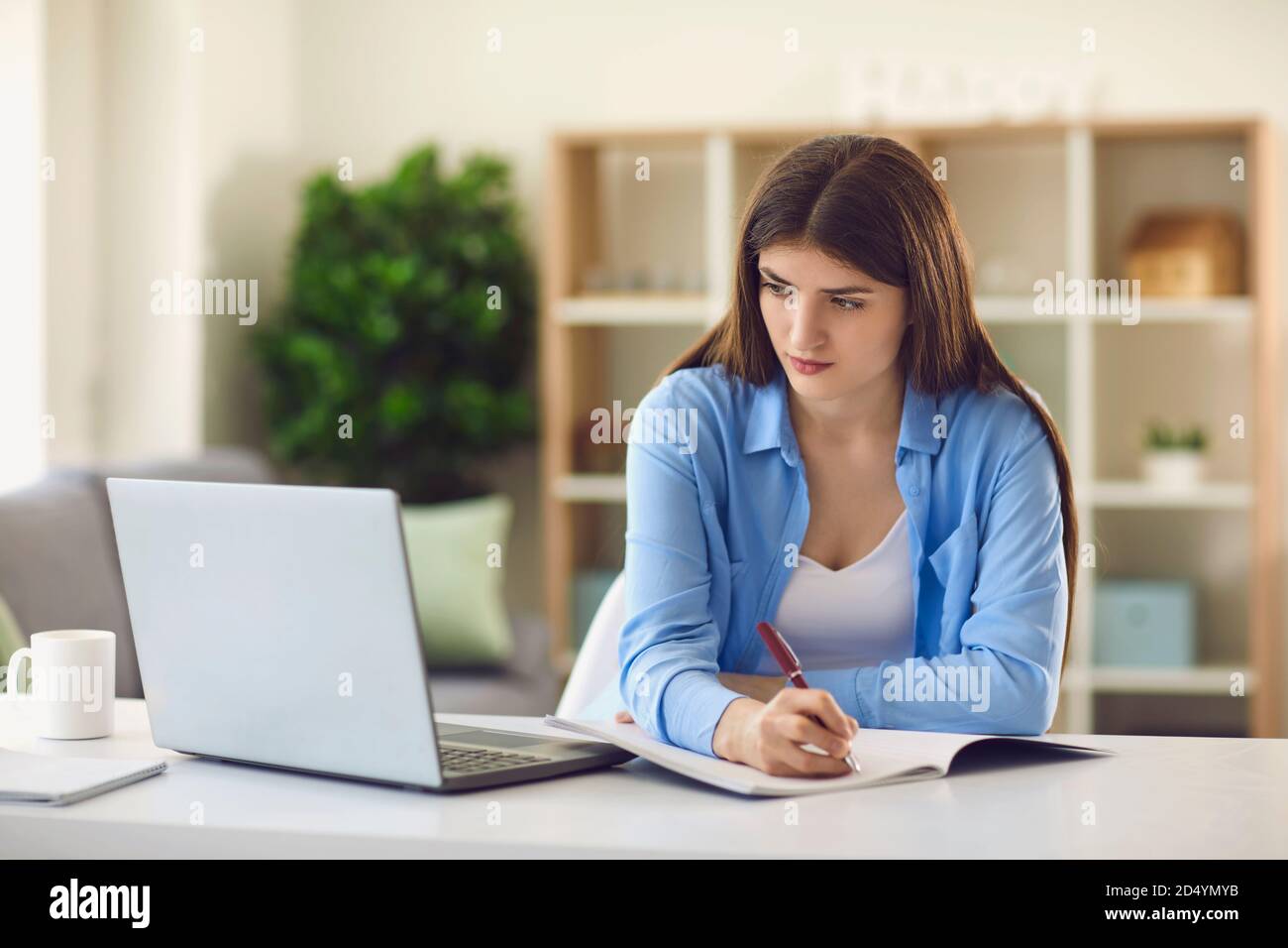 Une jeune étudiante écrit dans un ordinateur portable tout en regardant une conférence en ligne assis au bureau à la maison Banque D'Images