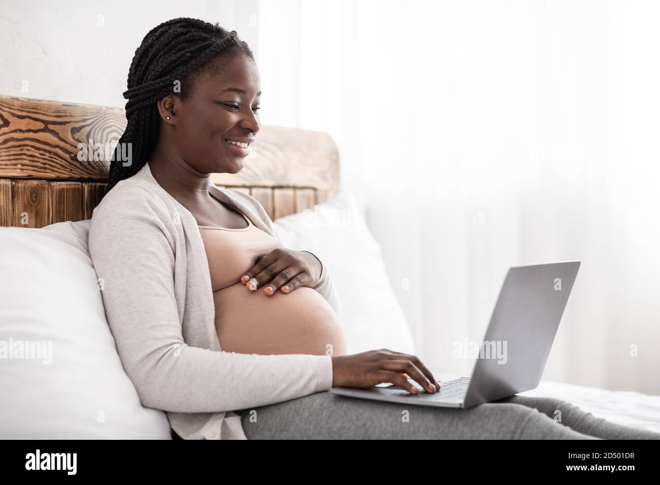 Charmante femme enceinte détente avec son ordinateur portable, de la navigation ou de discuter avec des amis en ligne, assis sur le lit, panorama avec l'espace vide Banque D'Images