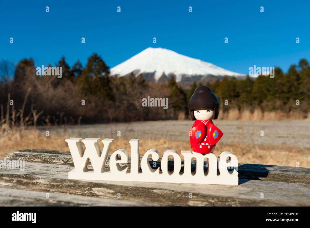 Magnifique poupée kokeshi traditionnelle japonaise vêtue de kimono rouge sur le mot 'Welcome'. Arrière-plan avec le Mont Fuji. Tir horizontal. Banque D'Images
