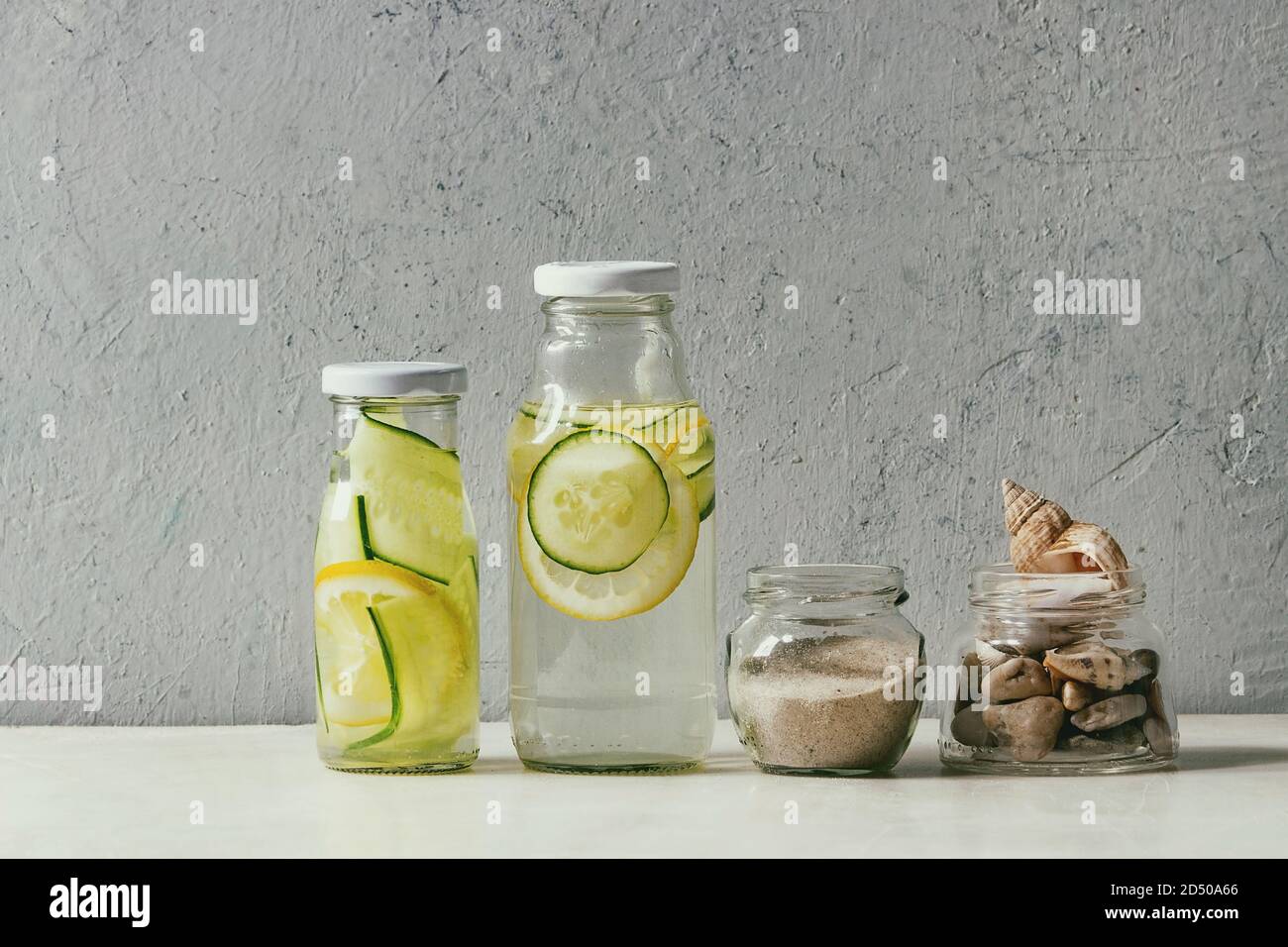 Thème d'été. Deux bouteilles en verre avec de l'eau de perfusion au citron et au concombre, des coquillages et du sable dans des pots en verre sur une table en marbre blanc, mur en béton à Banque D'Images